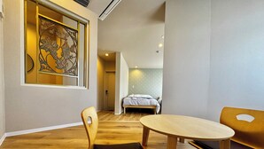 Quarto moderno com piso de madeira, mesa redonda, duas cadeiras, uma cama e um objeto decorativo na parede.