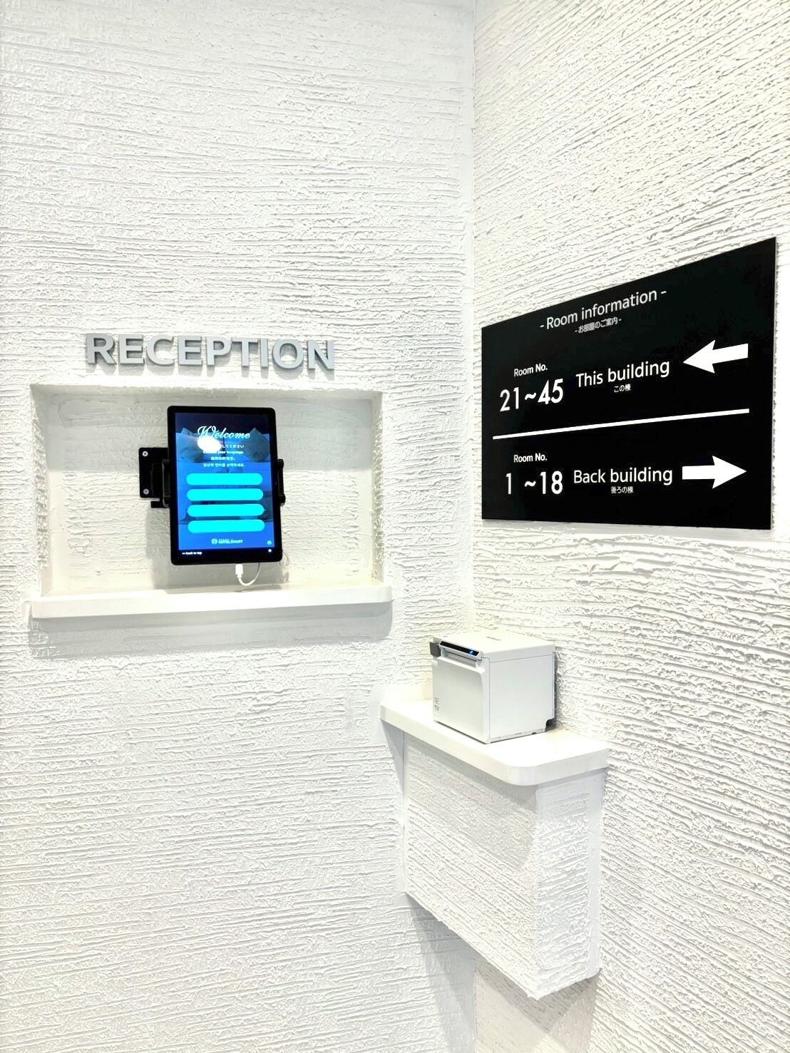A reception area with a digital screen displaying a welcome message, a sign indicating room numbers 21-45 and 1-18, and a directional sign for the back building.