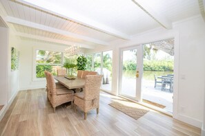 A dining area with a wooden table and wicker chairs, large windows, and a view of a deck and greenery. - Live Oak on The Indian River (Vero Beach)