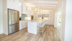 Una cocina moderna con armarios blancos, un refrigerador de acero inoxidable, una isla central con taburetes de bar, iluminación colgante y techo de madera.