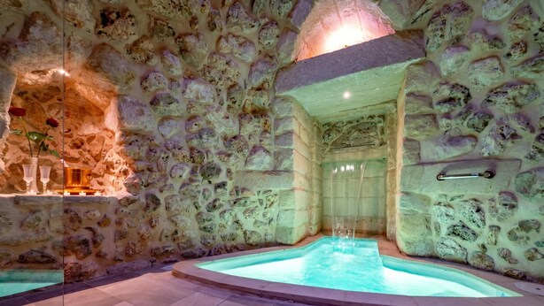 A stone-walled spa with a hot tub, a wooden shelf with vases, and a metal handrail.