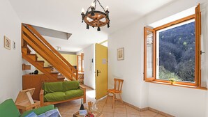 A spacious living area with a wooden staircase, a green sofa, a wooden coffee table with magazines, and a large window offering a view of a forested area.