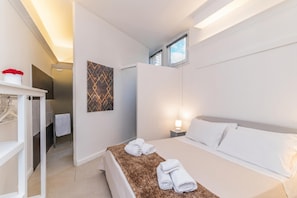 A hotel room with a bed, a bedside table, a wall-mounted lamp, a towel rack, and a door leading to a bathroom.