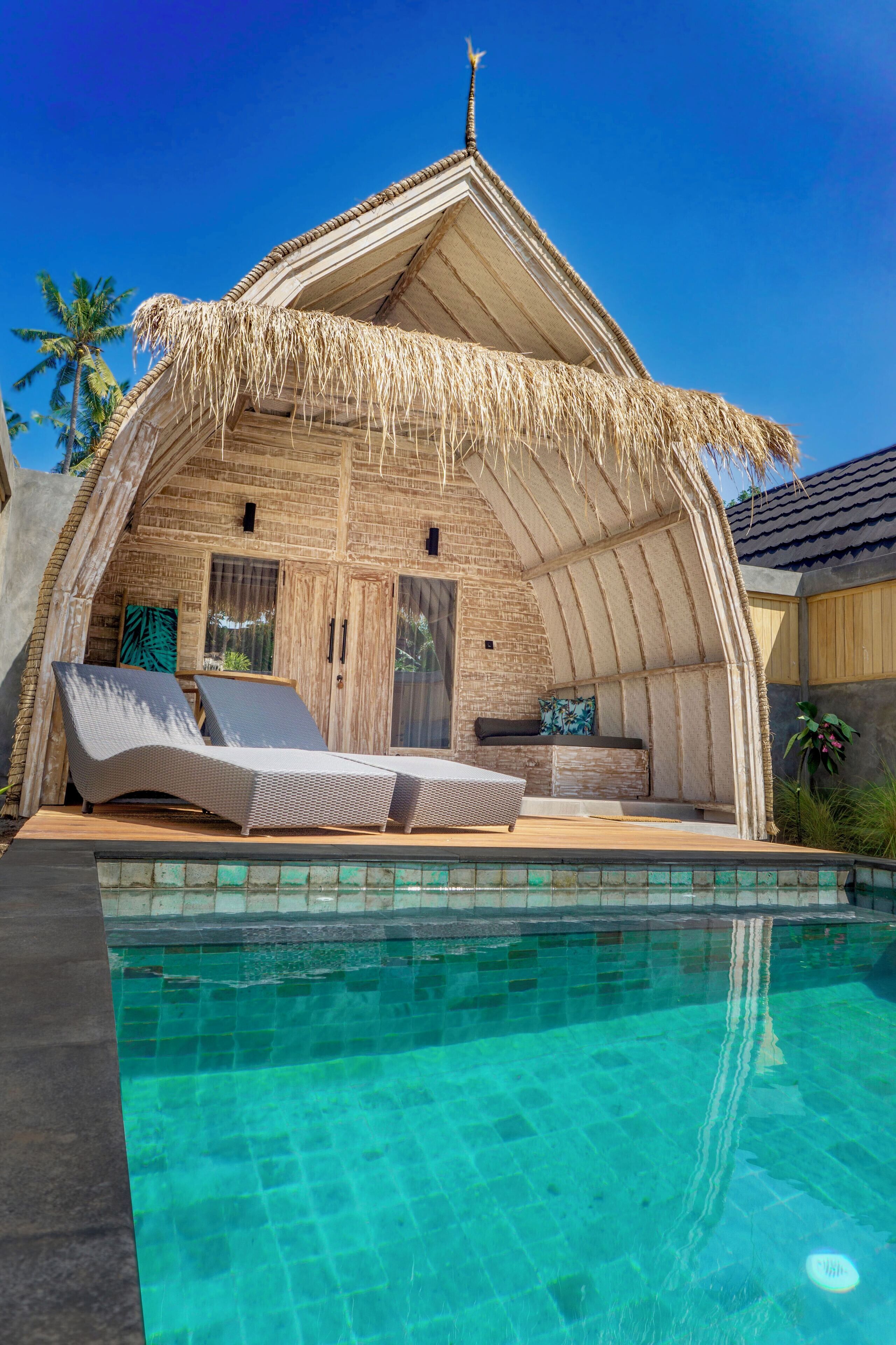 Un bungalow au toit de chaume, doté d’une piscine, de chaises longues et offrant une vue sur les palmiers.
