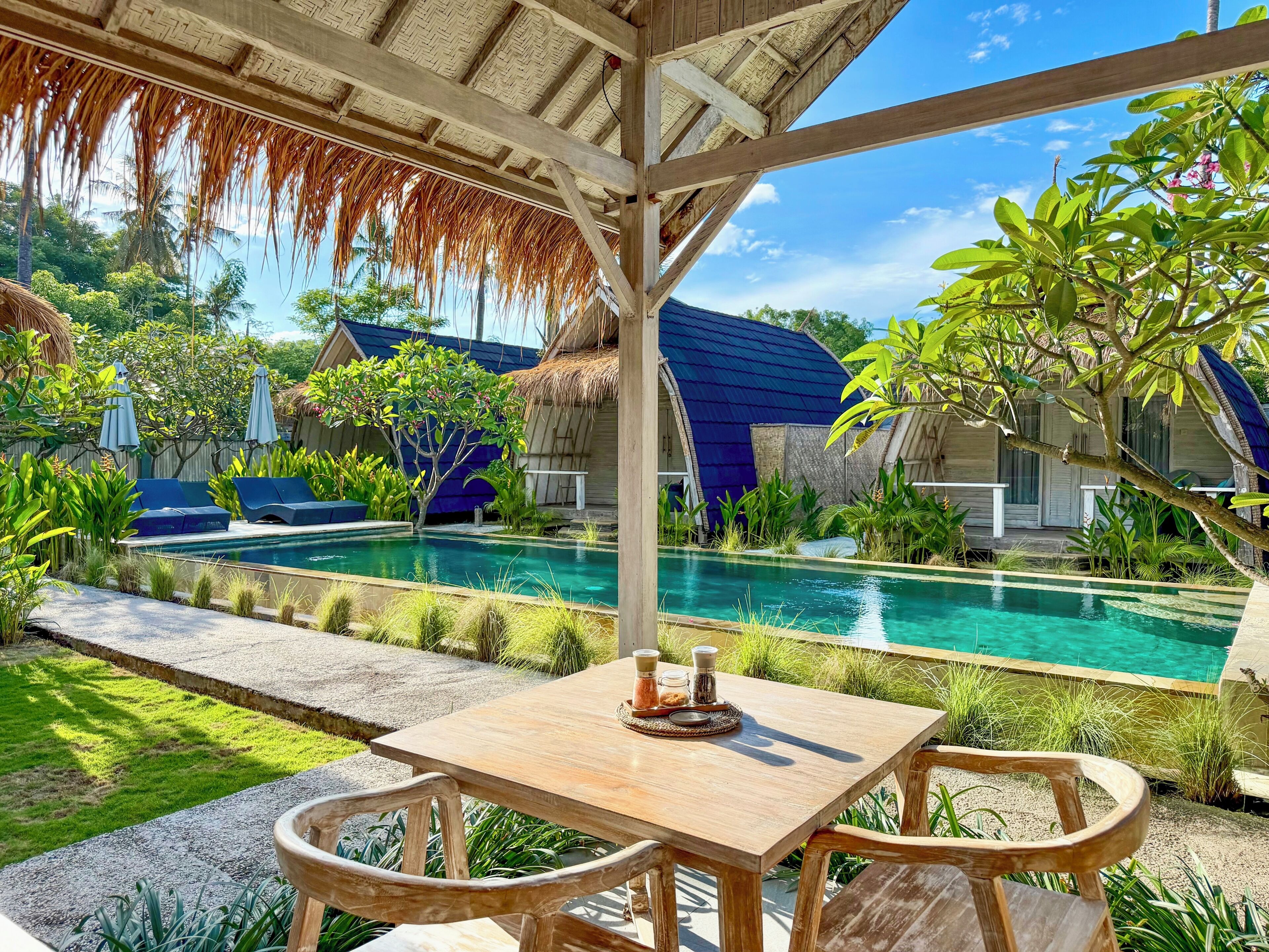 A poolside area with a wooden table and chairs, a thatched roof structure, and a view of a pool and overwater bungalows.
