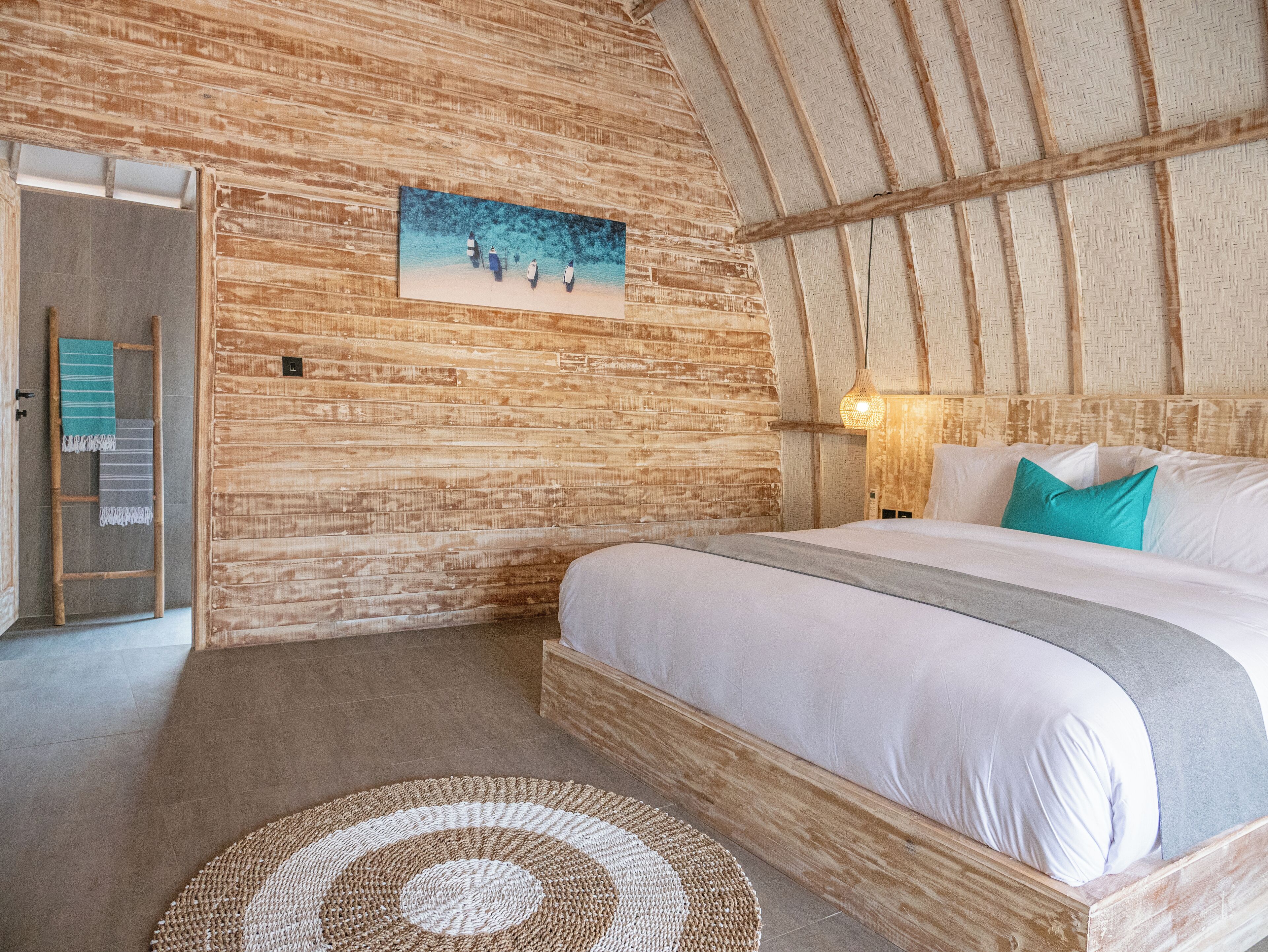 A cozy room with a wooden ceiling, a bed with white bedding, a painting on the wall, and a woven rug.