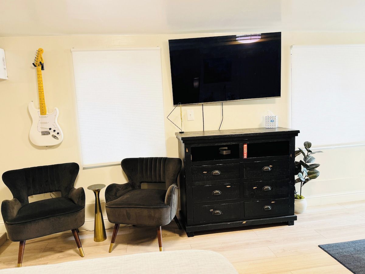 Una sala de estar con un mueble negro para centro de entretenimiento, dos sillas negras, una guitarra blanca y un televisor montado en la pared.