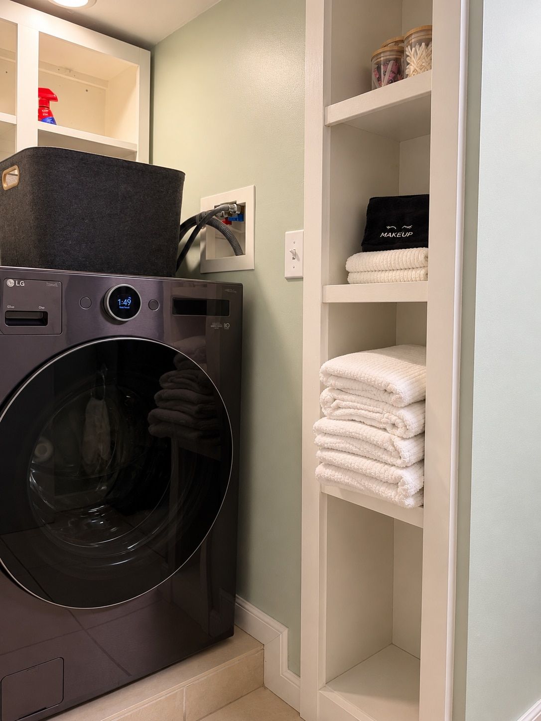 A laundry machine with a digital display, a white towel rack with folded towels, and a shelf with jars and a black bag labeled 'MAKEUP'.