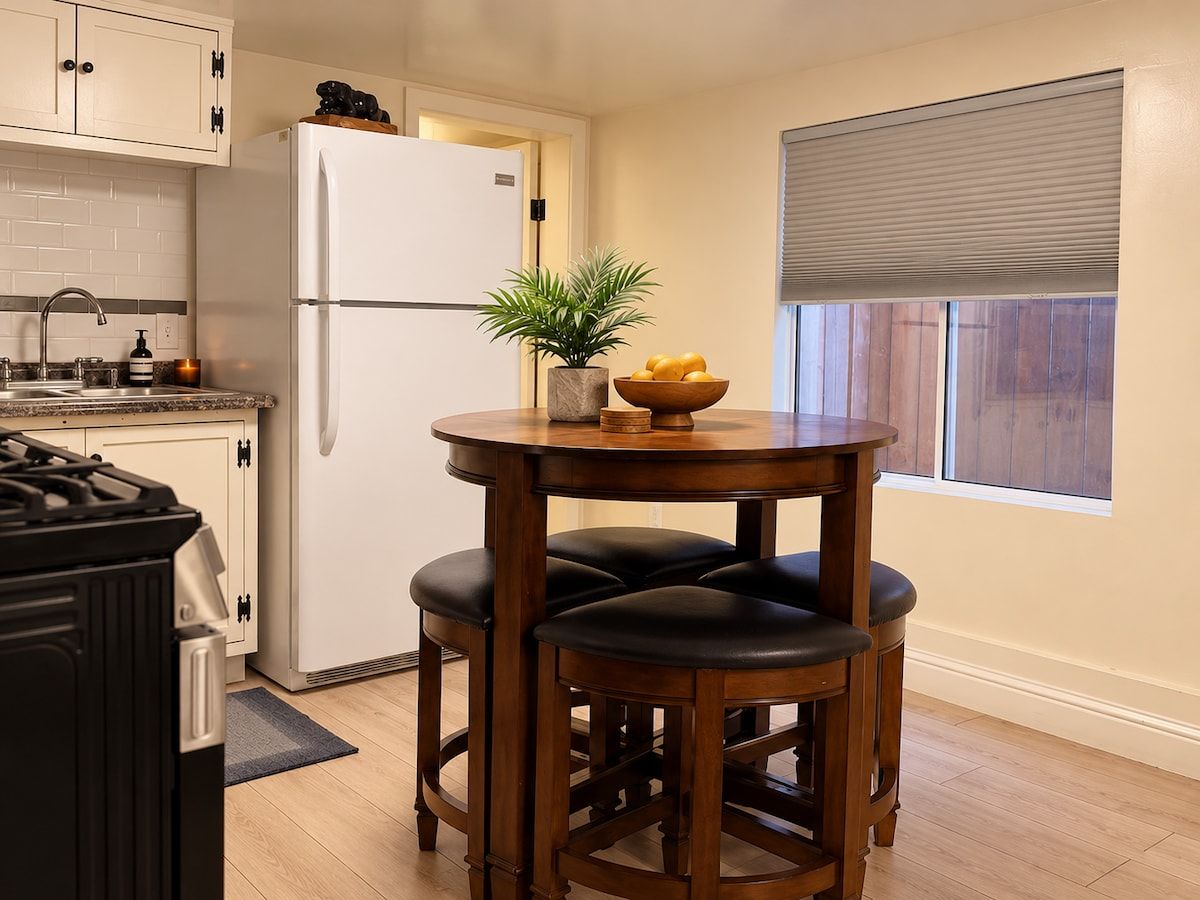 A kitchen with a white refrigerator, a wooden table with two chairs, a potted plant, and a window with blinds.
