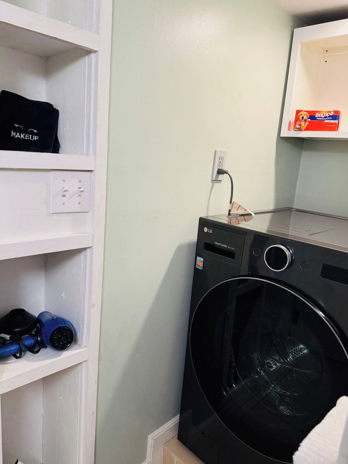 Un cuarto de lavado con una lavadora negra, un estante con un neceser y un anuncio de Megashirt en la pared.