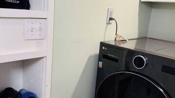 Un cuarto de lavado con una lavadora negra, un estante con un neceser y un anuncio de Megashirt en la pared.