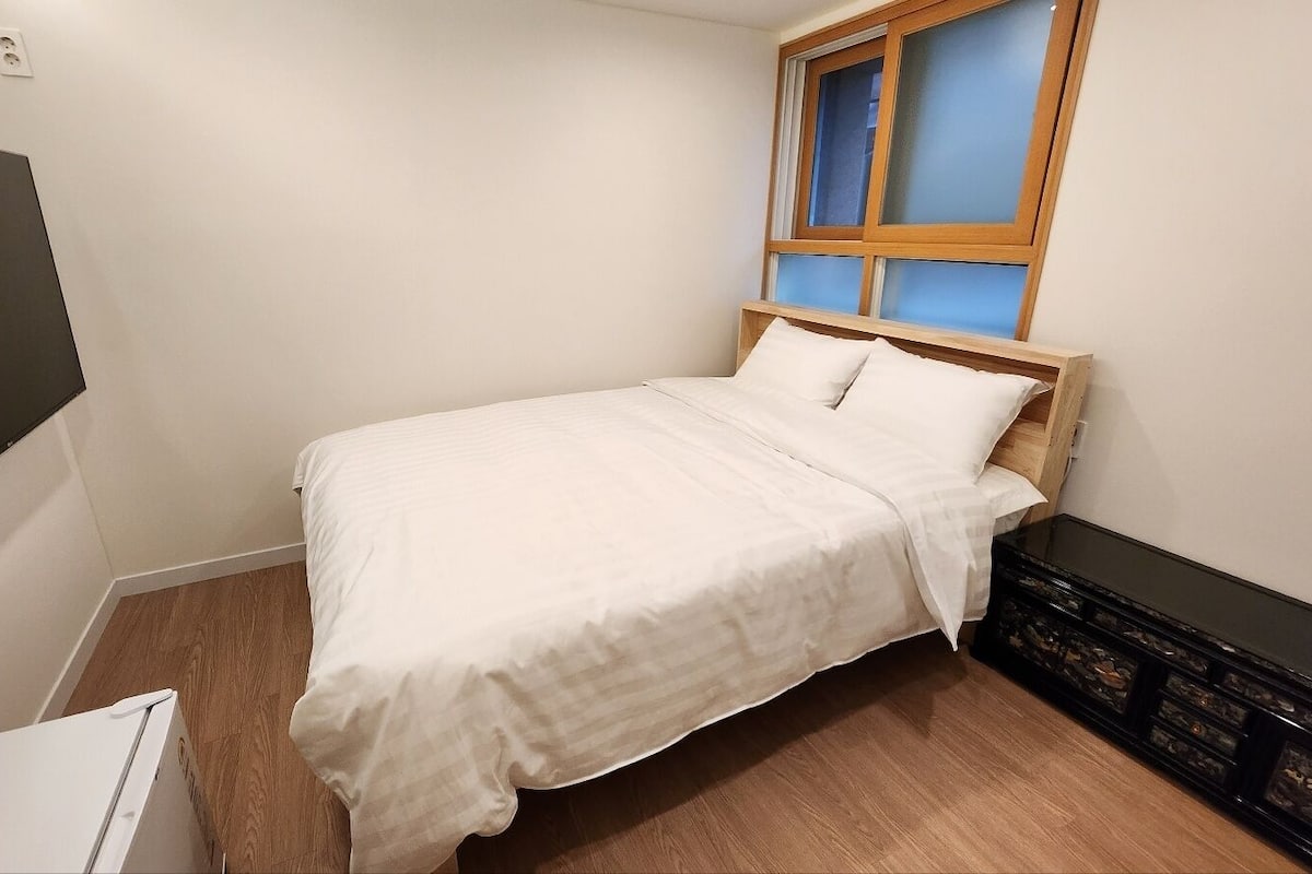 A small, neatly arranged bedroom with a single bed, a window with blue-tinted glass, and a black storage unit against the wall.