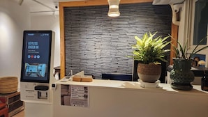 Una reception con una scrivania e uno schermo digitale che mostra 'KEYLESS CHECK IN', una parete in pietra e piante in vaso.