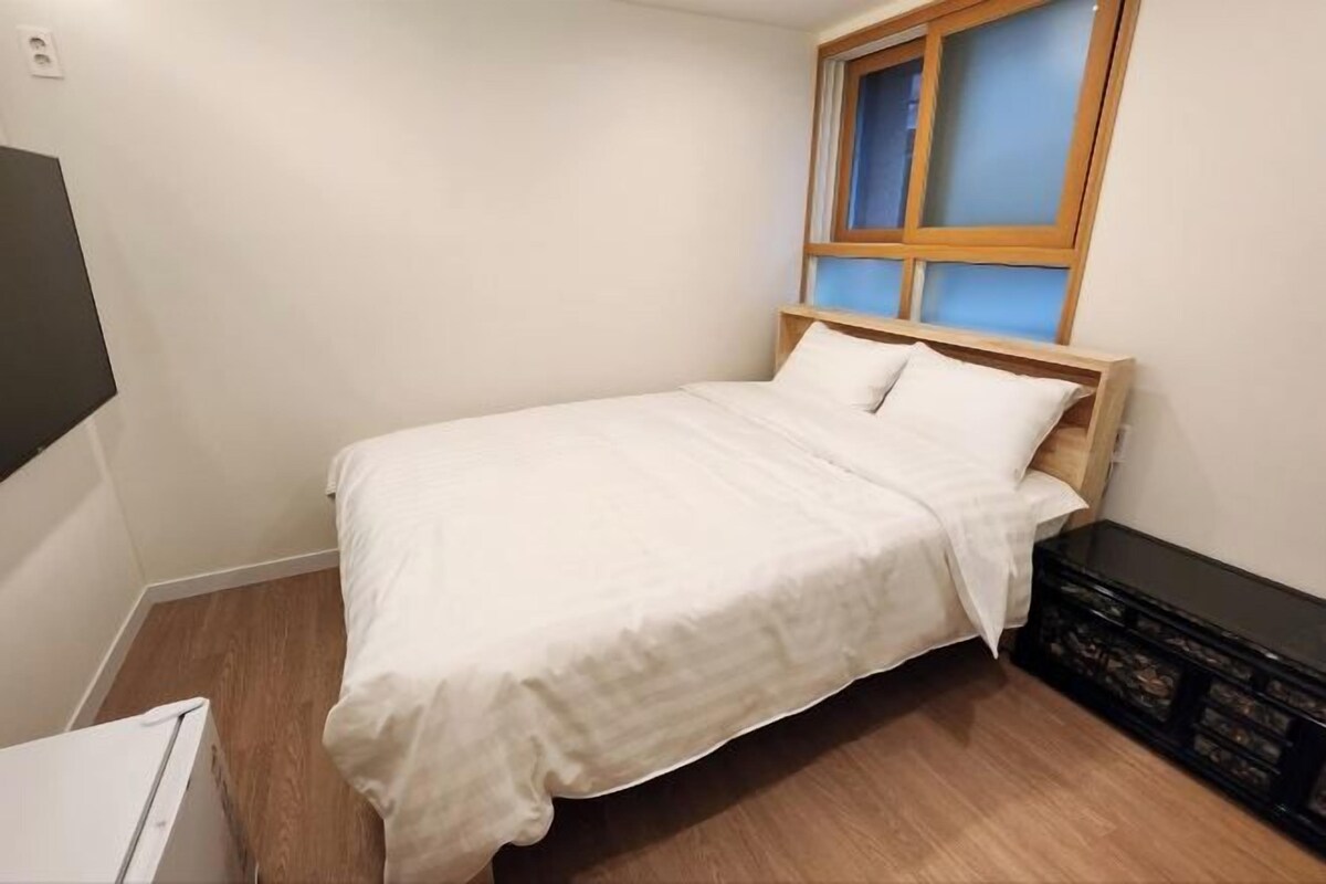 Un letto singolo con testiera in legno, un materasso bianco e due cuscini. Si vede una finestra con vetro azzurro e cornice in legno. Contro il muro, un mobile contenitore nero.