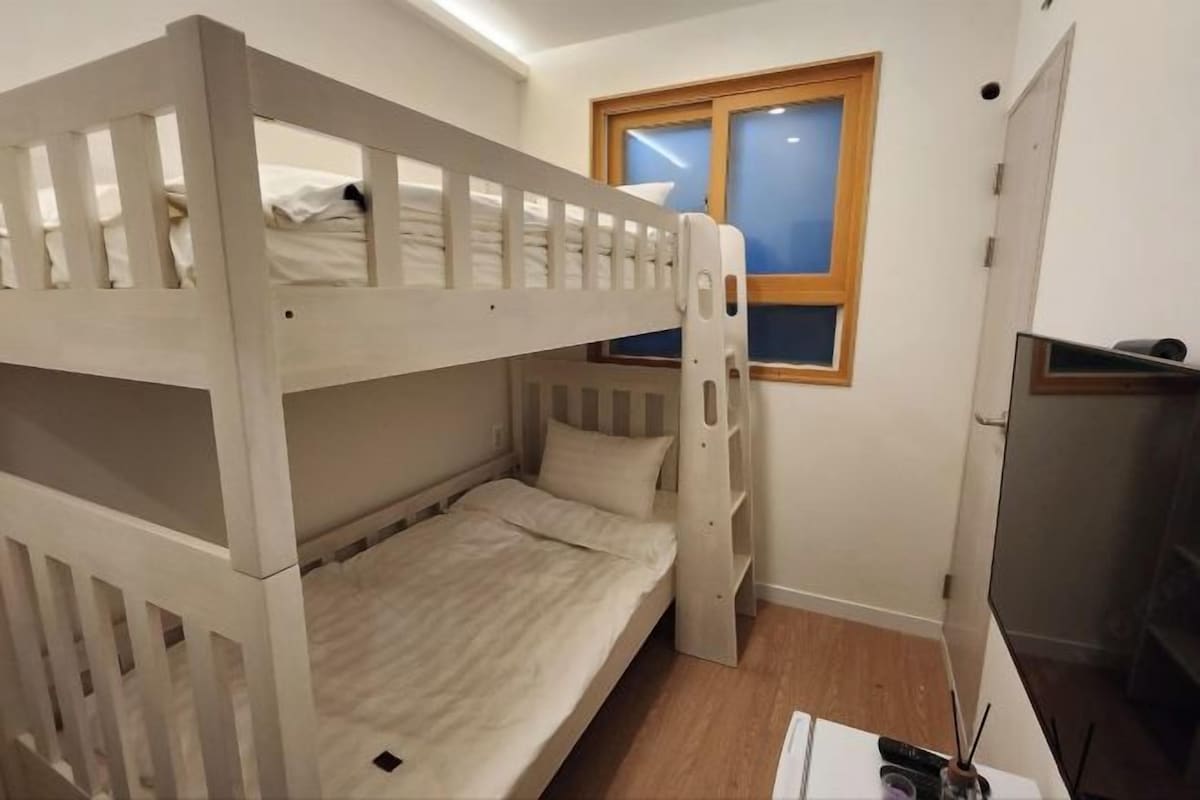 A bunk bed room with a window, a desk with a chair, and a TV.