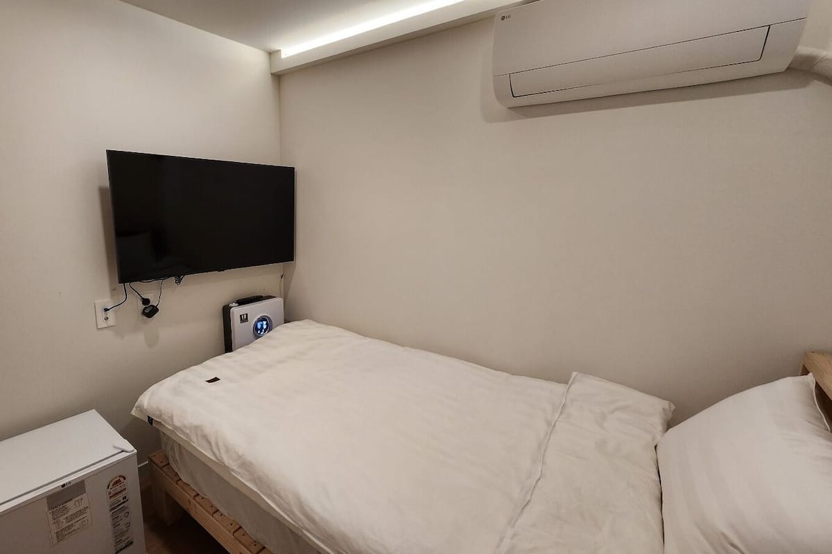 A small, clean room with a single bed, a TV mounted on the wall, a bedside table, and a wall-mounted air conditioner.