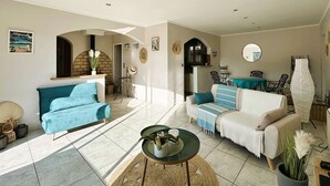 A spacious living area with a blue sofa, a white sofa, and a round coffee table. There is a fireplace with a brick surround, a dining area with a table and chairs, and a kitchen visible through an archway.