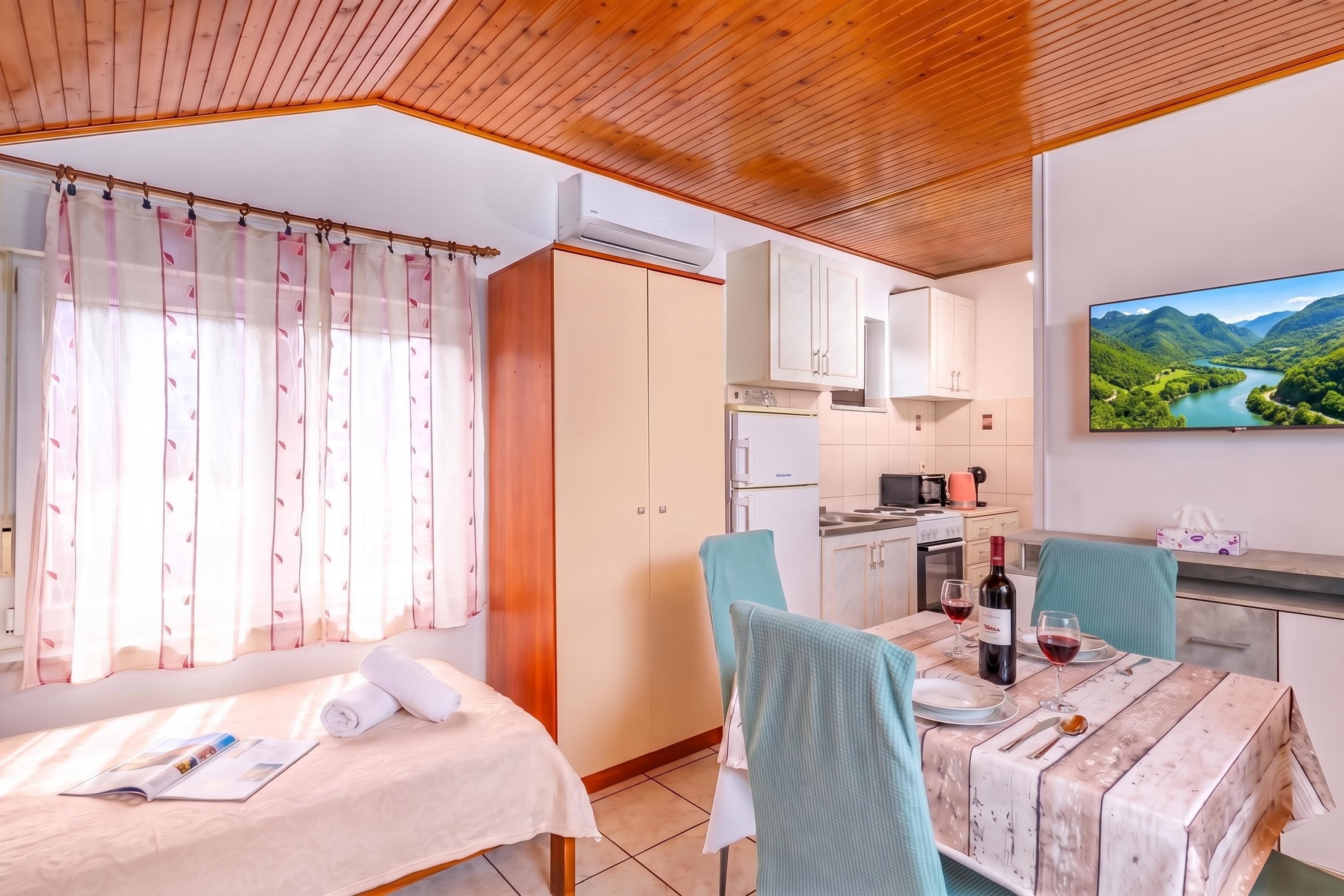 A room with a bed, a dining table with chairs, a kitchen area, and a television displaying a scenic landscape.
