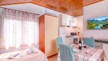 A room with a bed, a dining table with chairs, a kitchen area, and a television displaying a scenic landscape.
