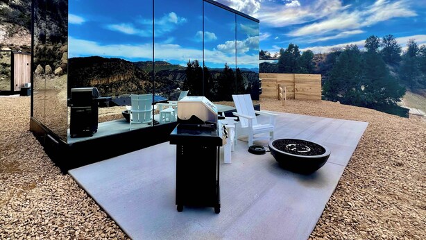Un espace extérieur moderne, aménagé avec des sièges, doté d’un mur de verre qui reflète un paysage montagneux, d’un foyer et de chaises blanches.