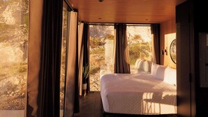 Une chambre à coucher avec un lit, une table, une chaise, une fenêtre avec des rideaux et une vue sur l’extérieur.