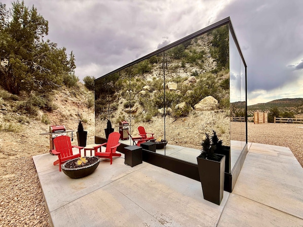 A modern outdoor seating area with red chairs, a grill, and a large mirror reflecting the surrounding landscape.