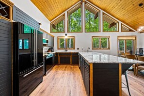 A modern kitchen with a large island, wooden flooring, and large windows offering views of the forest.