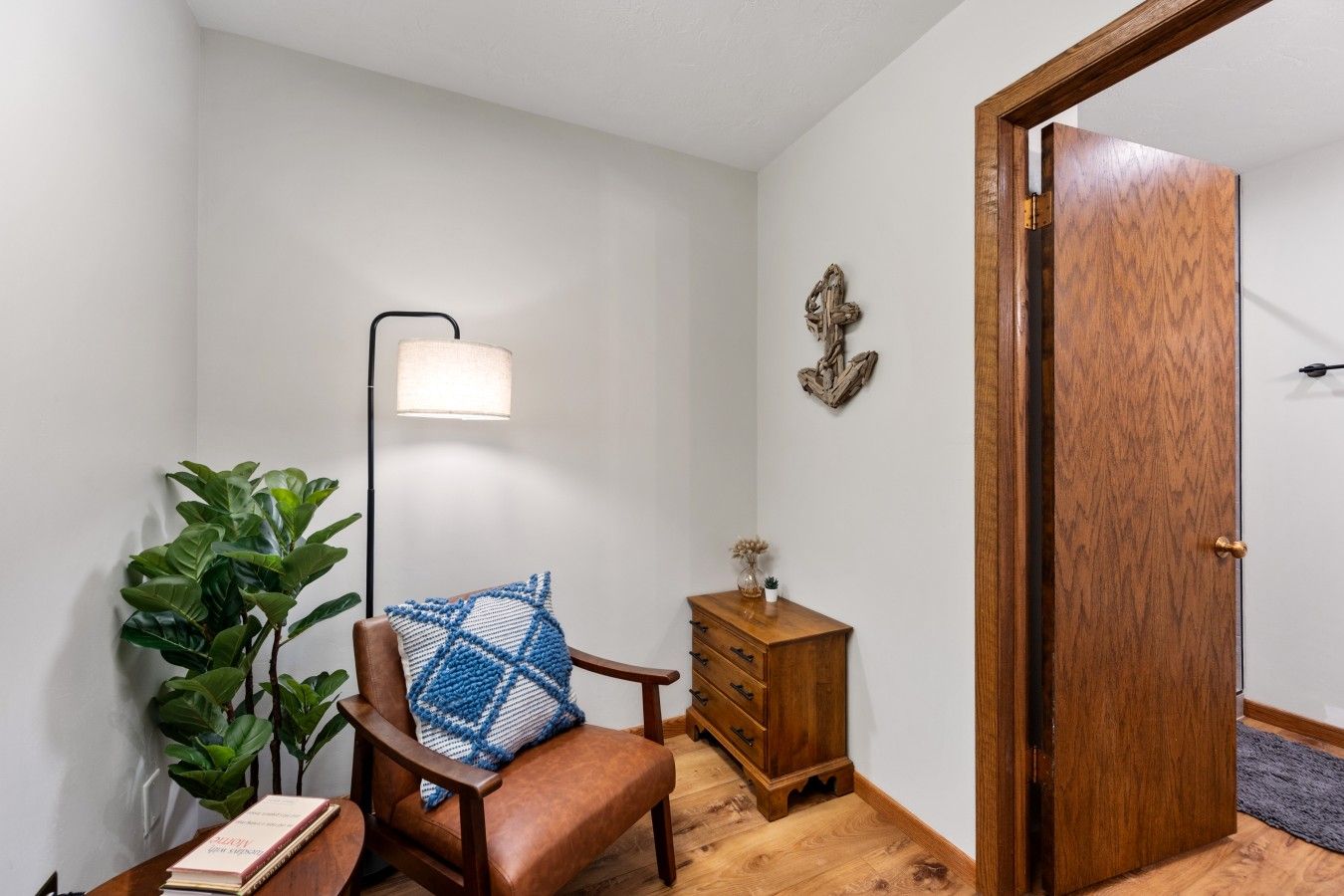 A room with a wooden floor, a brown leather chair with a blue cushion, a small wooden dresser, a floor lamp, and a potted plant.