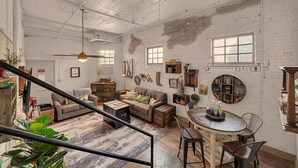 A spacious loft with exposed brick walls, a loft bed, a dining area with a round table, and a staircase. - Waco Memory Lane, Downtown Luxury Loft- Steps to Silos/Baylor/McLane (Waco)
