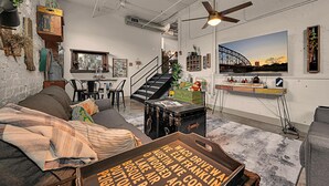 A cozy living room with a sofa, a dining table, and a staircase leading to a loft area. - Waco Memory Lane, Downtown Luxury Loft- Steps to Silos/Baylor/McLane (Waco)