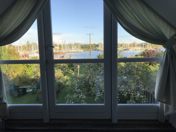 Een kamer met een groot raam met uitzicht op een jachthaven met boten en een tuin met bloeiende planten.