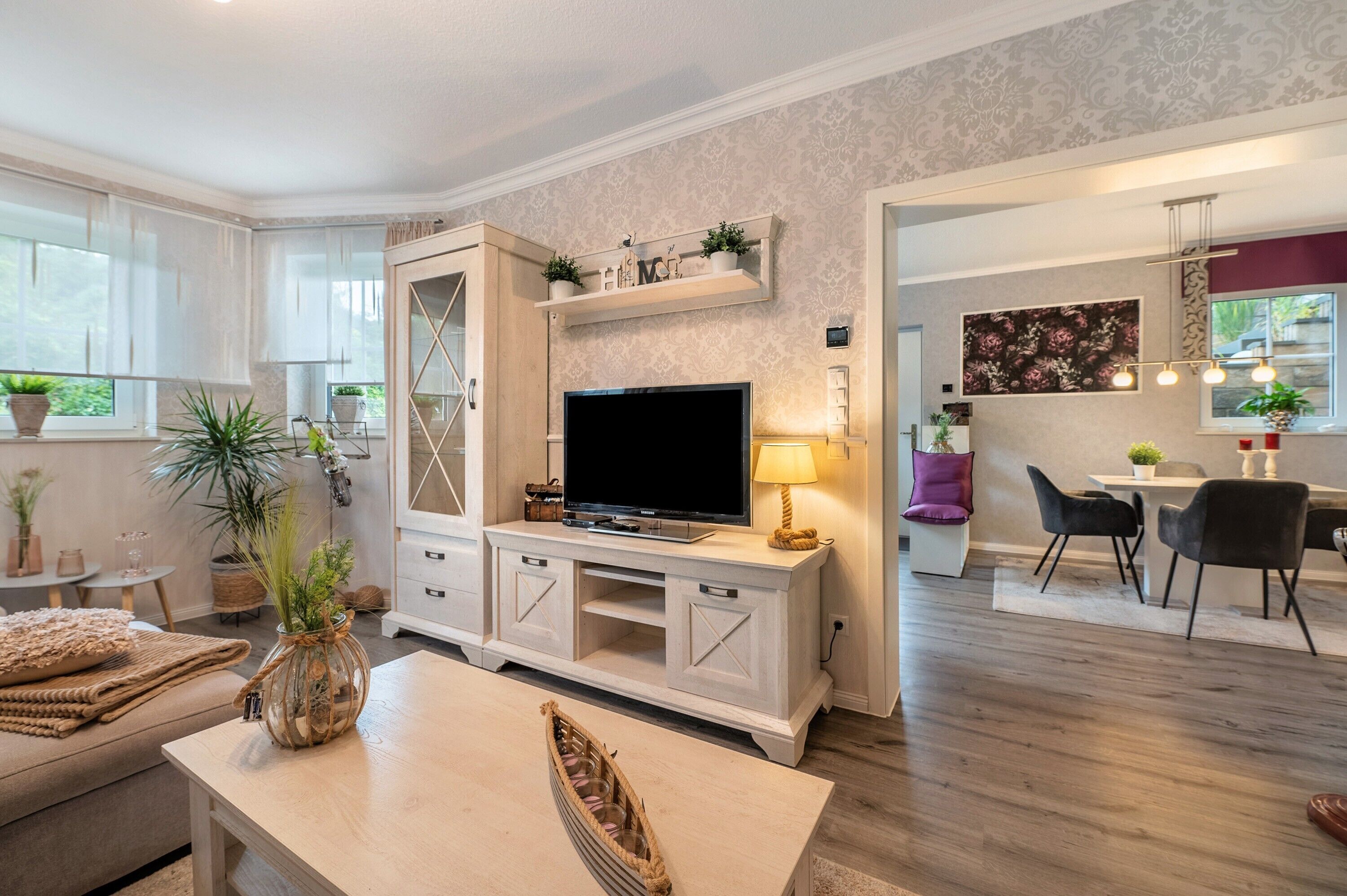 Un salon moderne avec un téléviseur, des meubles blancs, un coin repas et une cuisine visible au fond.