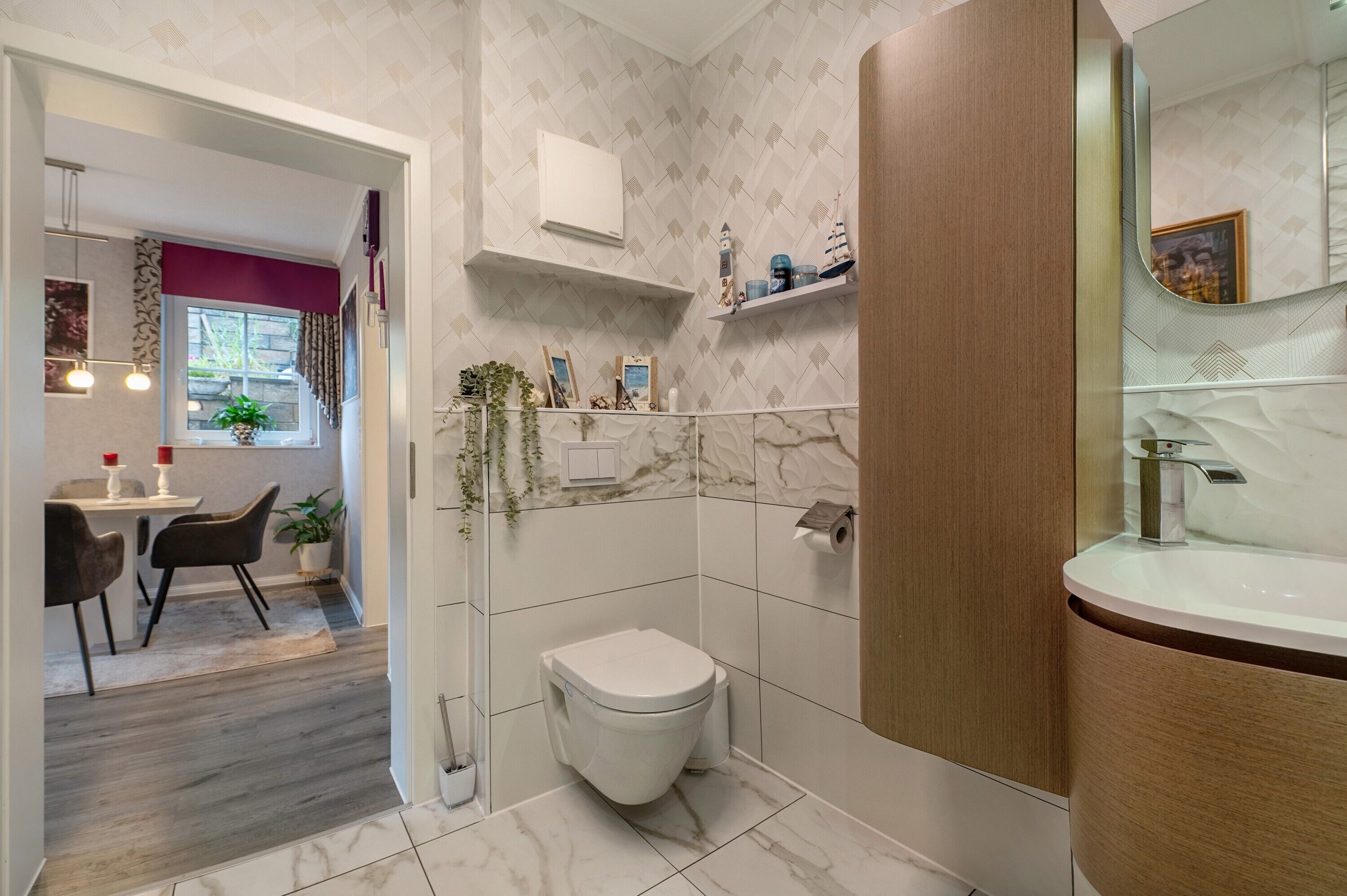 Un baño moderno con inodoro, lavabo y zona de ducha. Hay una ventana con cortinas y una pequeña mesa con sillas en la habitación contigua.