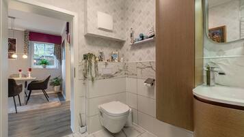 Un baño moderno con inodoro, lavabo y zona de ducha. Hay una ventana con cortinas y una pequeña mesa con sillas en la habitación contigua.