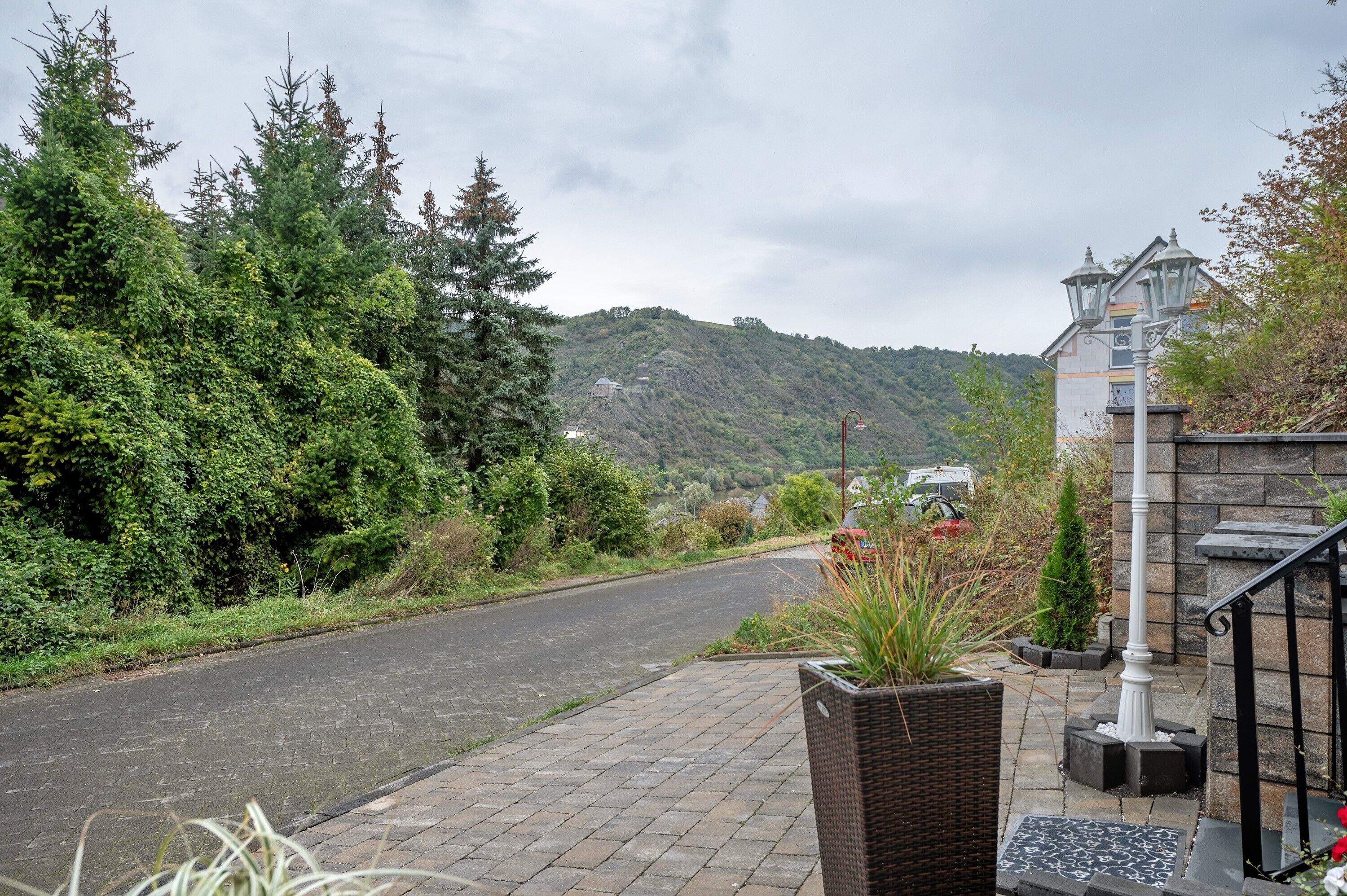 A paved pathway leading to a building with a traditional architectural style, surrounded by lush greenery and a hilly landscape in the background.