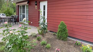 Een rode houten woning met een terras, een kleine tuin met bloemen en een auto op de oprit.