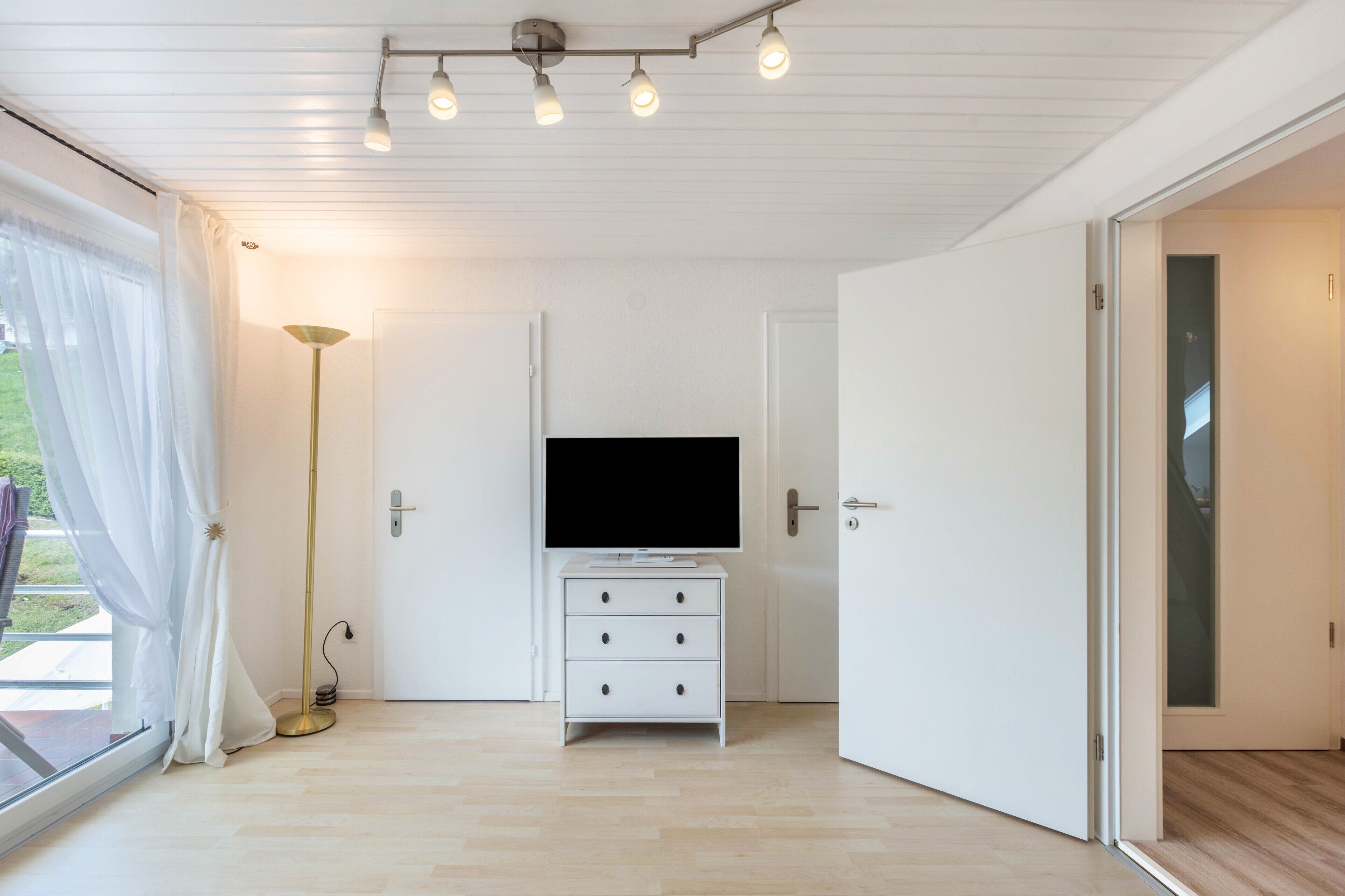 A modern room with a flat-screen TV on a white dresser, a floor lamp, and a sliding glass door leading outside.