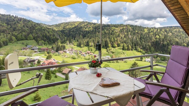 A balcony with a yellow umbrella, a table with a cushion, a chair, and a scenic view of a forested valley.