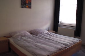 Una cama individual con estructura de madera, una mesita de noche, una lámpara fijada a la pared y una ventana con cortinas.