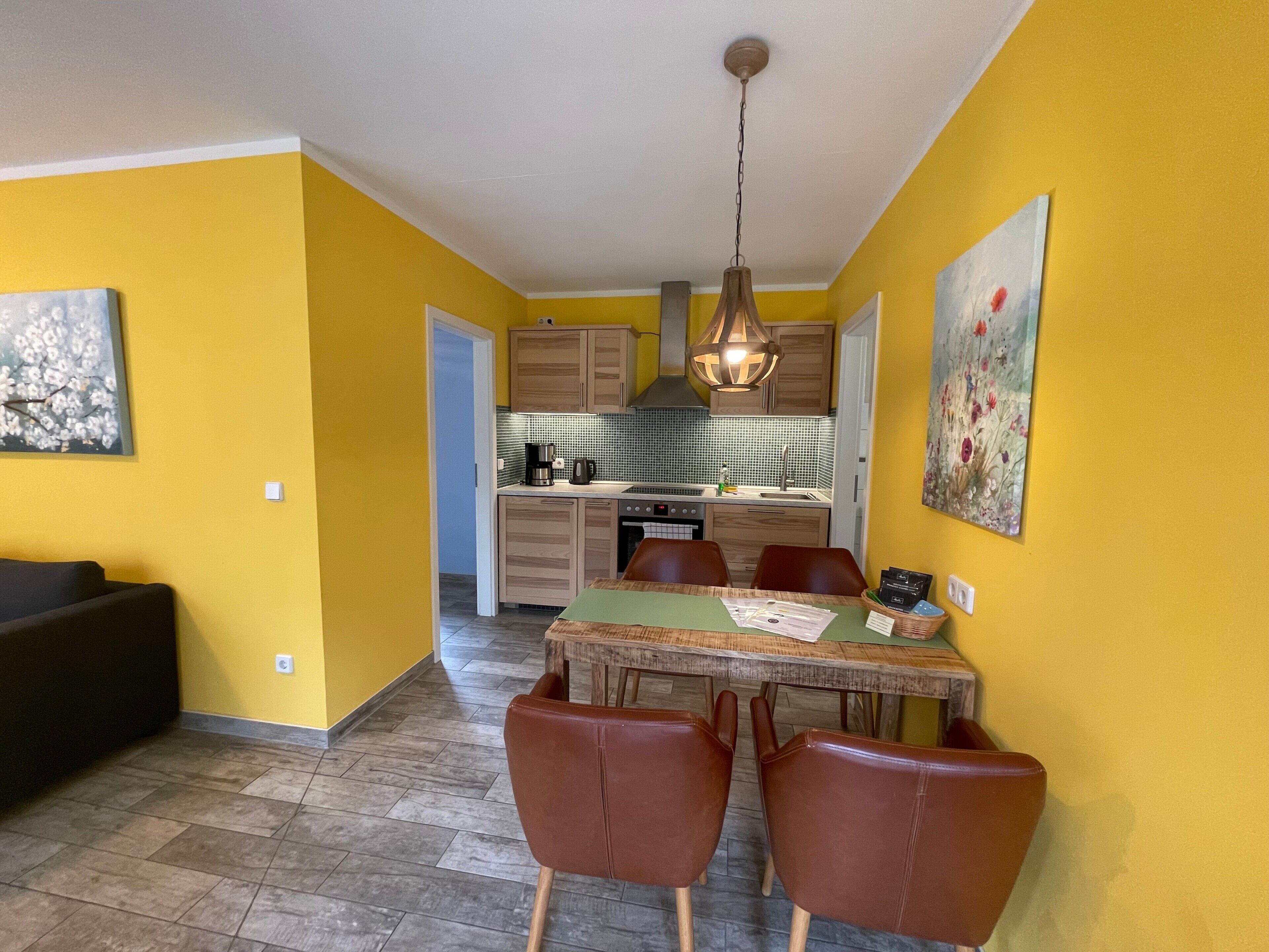 A dining area with a table and chairs, a kitchen in the background, and a yellow wall.