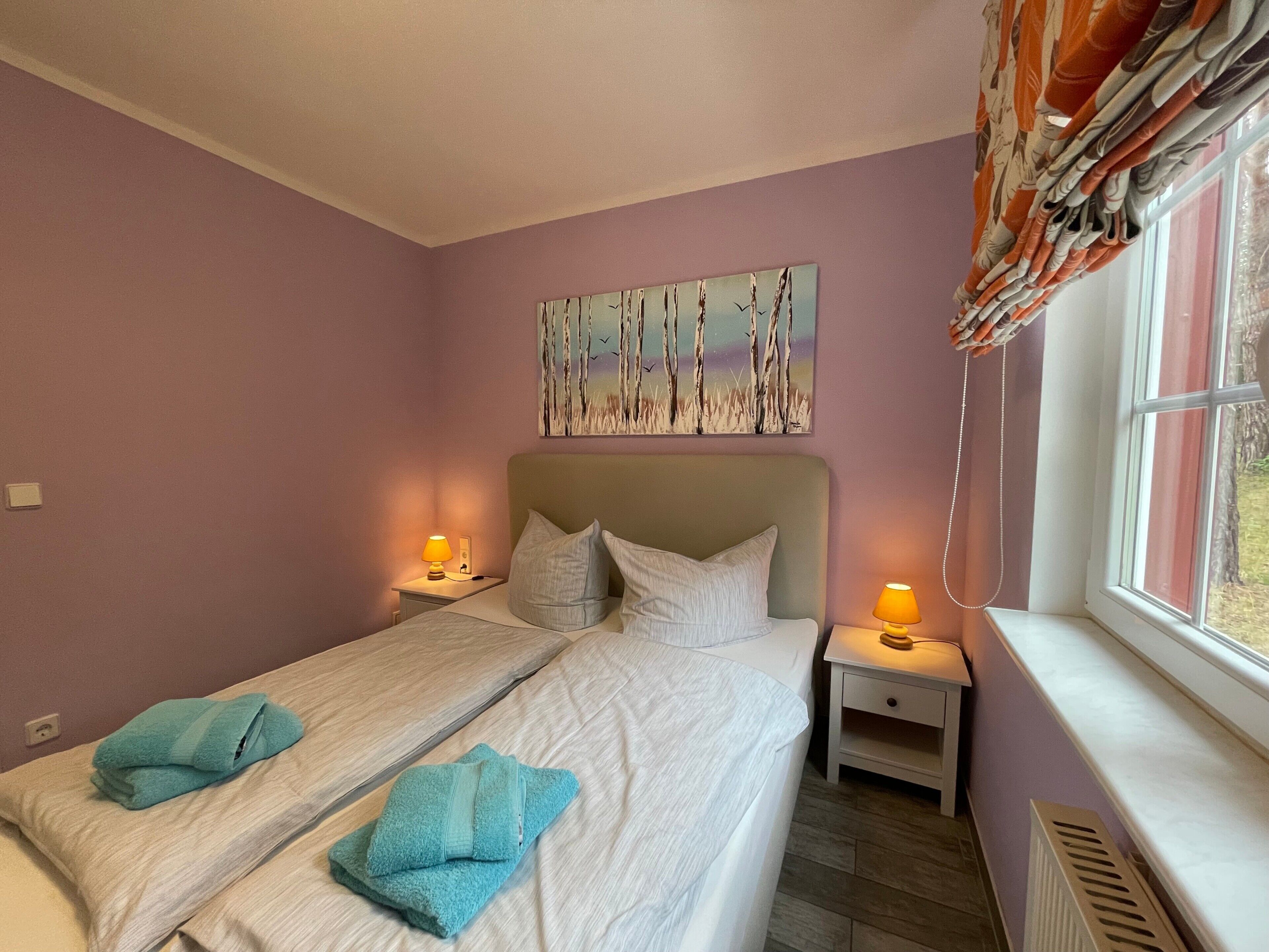 A bedroom with two beds, a nightstand with lamps, a painting on the wall, and a window with curtains.