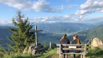 Zwei Personen sitzen auf einer Holzbank und blicken auf eine gebirgige Landschaft. Ein Wegweiser zeigt auf einen Aussichtspunkt.