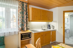 A compact kitchen with wooden cabinets, a microwave, a sink, and a window with curtains. - Studio Apartment 'Appartement Haus Voglreiter' with Mountain View, Shared Garden and Wi-Fi (Kaprun)