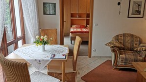 Una habitación con una mesa de comedor, sillas de mimbre, un sofá, una cama con colcha roja y blanca, y un armario de madera.