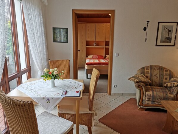 A room with a dining table, wicker chairs, a sofa, a bed with a red and white bedspread, and a wooden wardrobe.