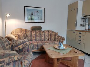 A living room with a patterned sofa, a wooden coffee table, a framed picture on the wall, and a kitchen area with cabinets and utensils.