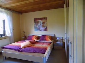 A bedroom with a bed, a nightstand, a lamp, a painting on the wall, and a window with curtains.