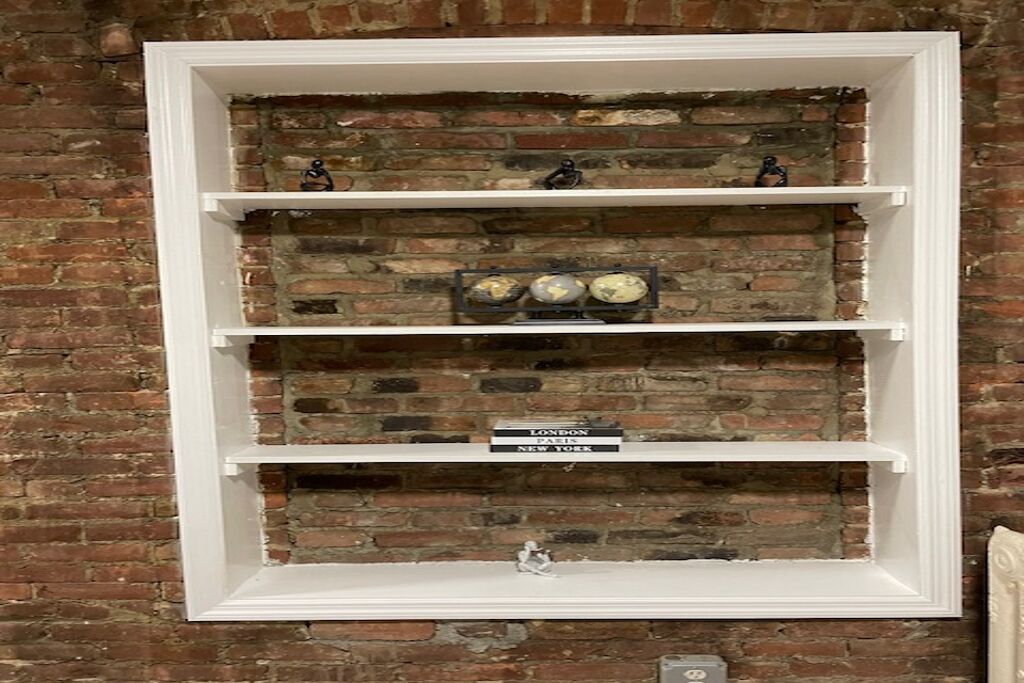A white-framed shelf with three levels against a brick wall. The top shelf holds decorative items, the middle shelf has globes, and the bottom shelf has a sign with city names.