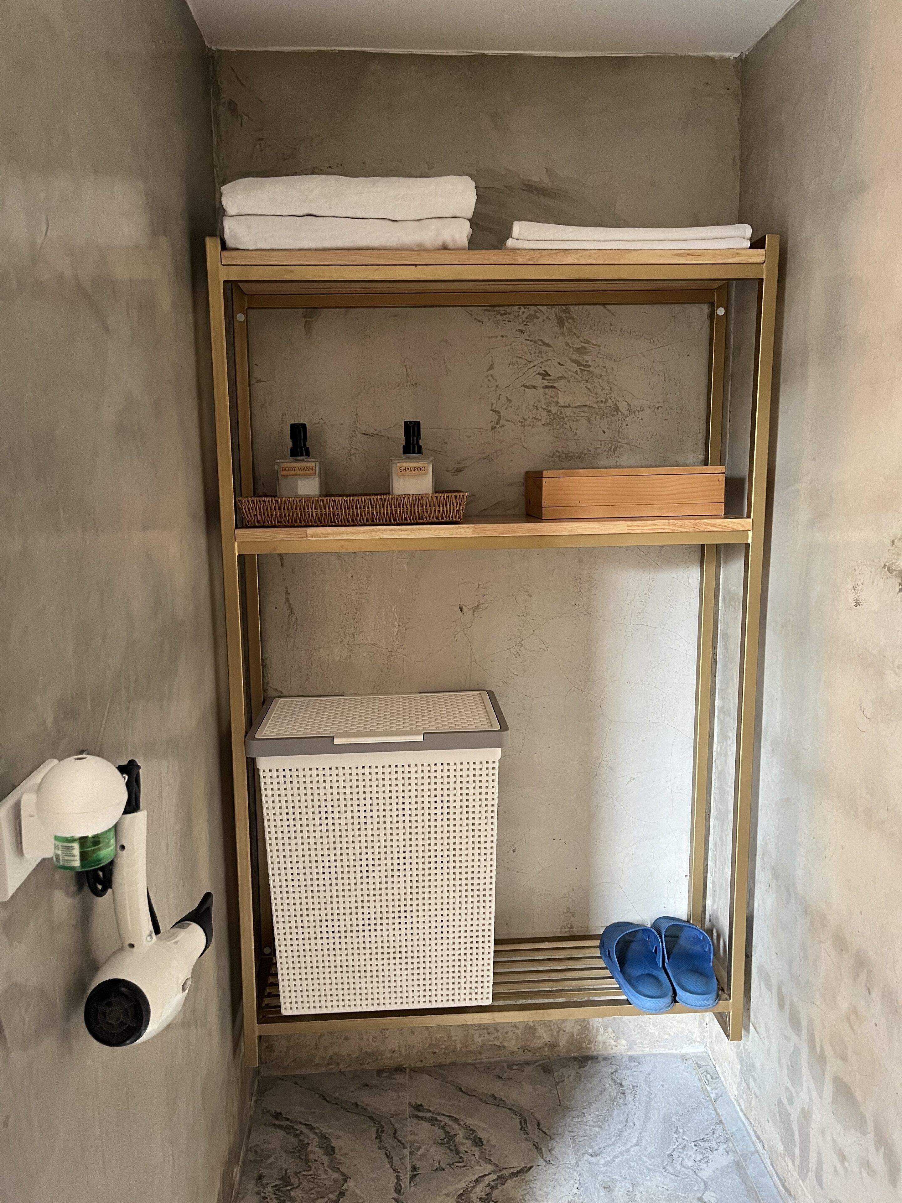 Une salle de bain avec une étagère en bois où sont posées des serviettes et des articles de toilette, un panier blanc et une paire de pantoufles bleues sur un porte-chaussures.