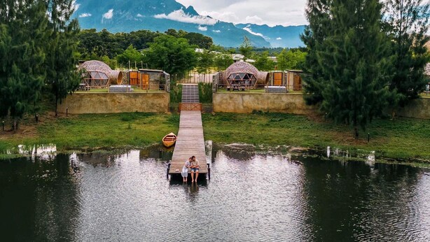 Un muelle de madera que conduce a una casa con estructura en forma de cúpula, rodeada de vegetación y una masa de agua.